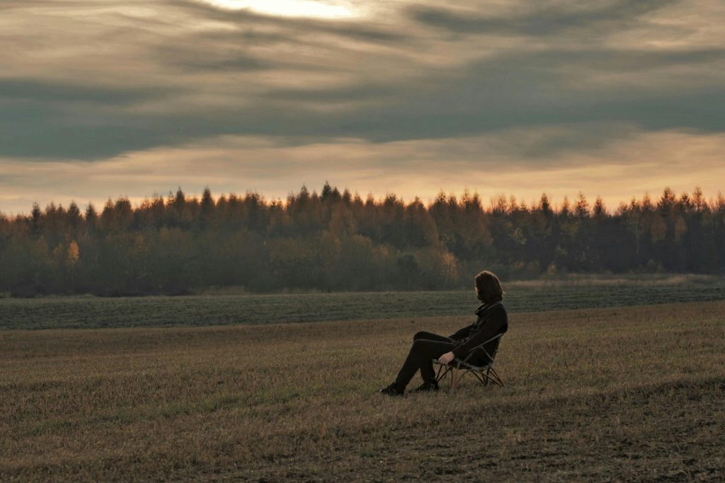 A solitary person sits on a chair in a tranquil countryside field at sunset, evoking peace. Une personne solitaire assise sur une chaise dans un champ paisible à la campagne au coucher du soleil, évoquant la paix.