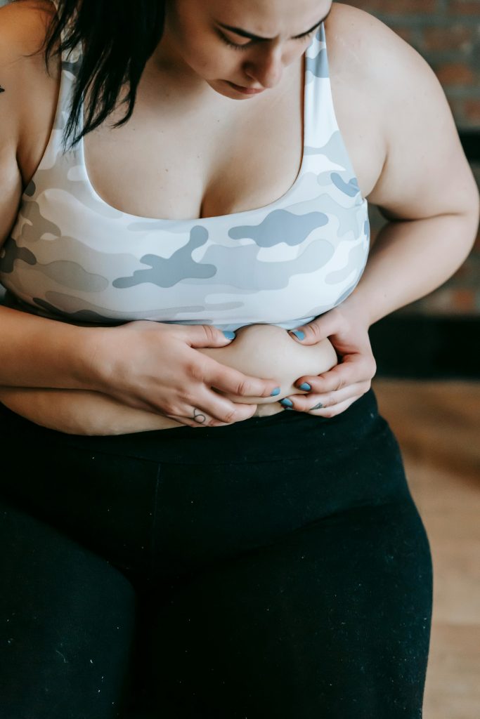 Crop sérieuse jeune femme forte en brassière de sport et legging touchant sa graisse abdominale pendant un entraînement en salle de sport