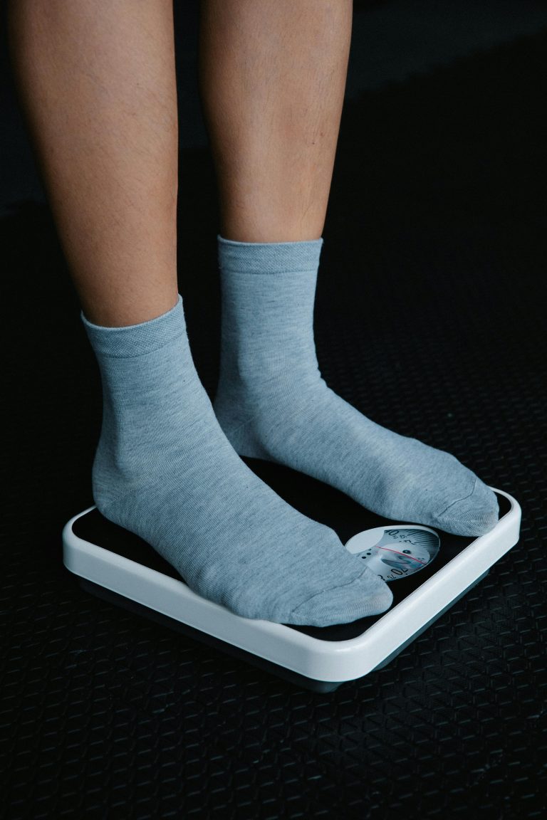 Close-up de pieds en chaussettes grises sur un pèse-personne posé sur un sol sombre.