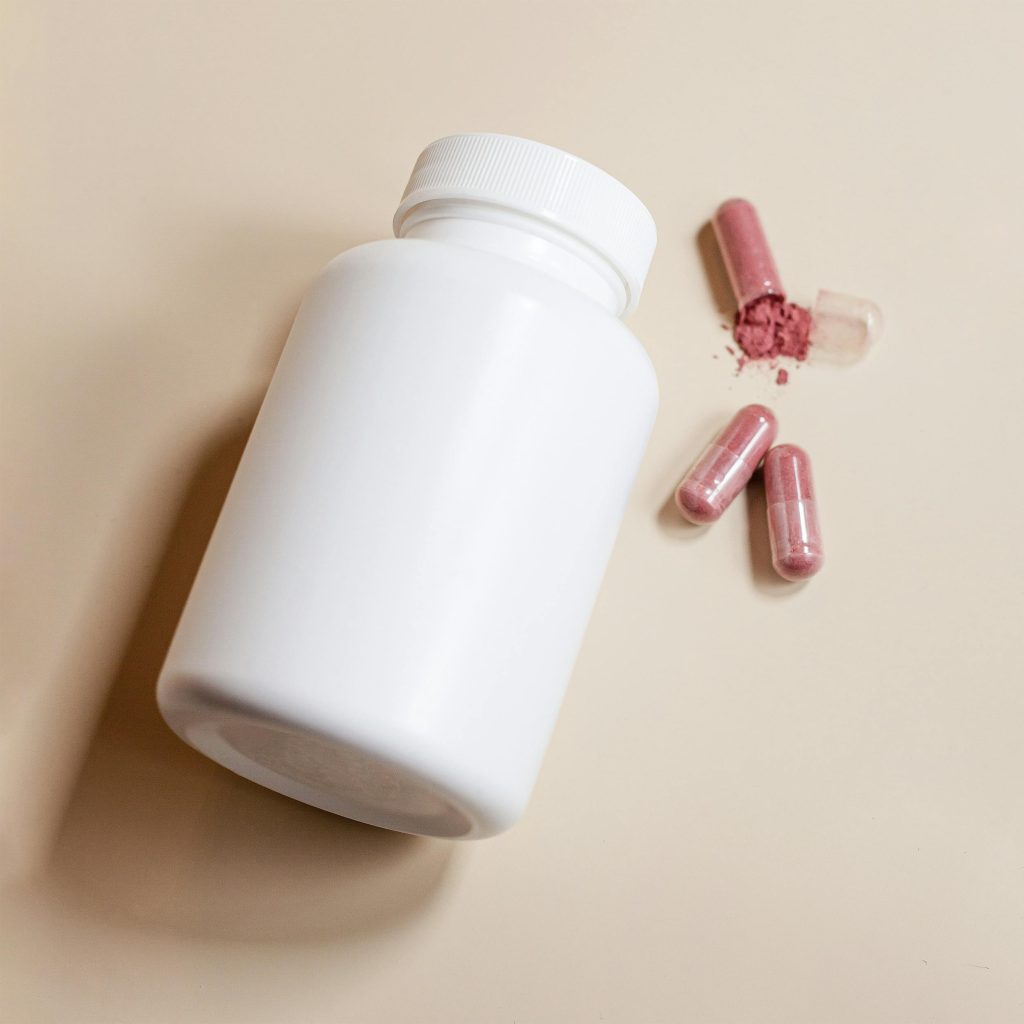 White pill bottle with spilled pink capsules containing powder, shot on a neutral background. Flacon de pilules blanc avec des gélules roses renversées contenant de la poudre, photographié sur un fond neutre.