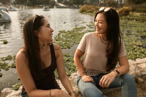 Two women sitting and smiling by the lake, enjoying the outdoors and friendship. Deux femmes assises et souriantes au bord du lac, profitant de l'extérieur et de l'amitié.
