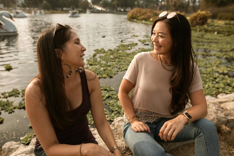 Two women sitting and smiling by the lake, enjoying the outdoors and friendship. Deux femmes assises et souriantes au bord du lac, profitant de l'extérieur et de l'amitié.