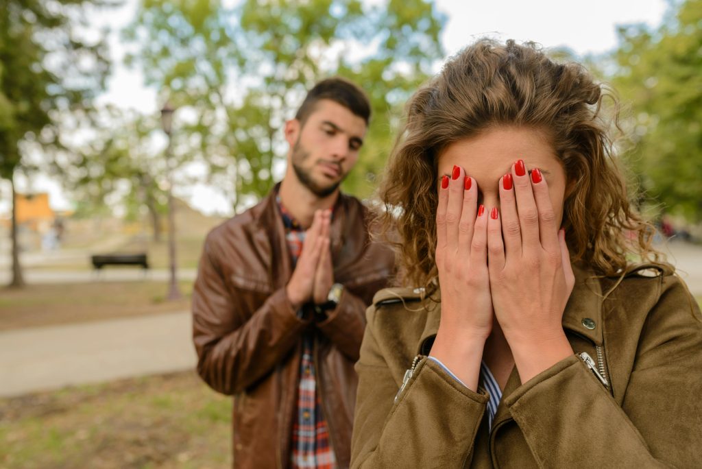 A young man apologizes while a woman covers her face, set in an outdoor park. Un jeune homme s'excuse tandis qu'une femme se couvre le visage, dans un parc extérieur.