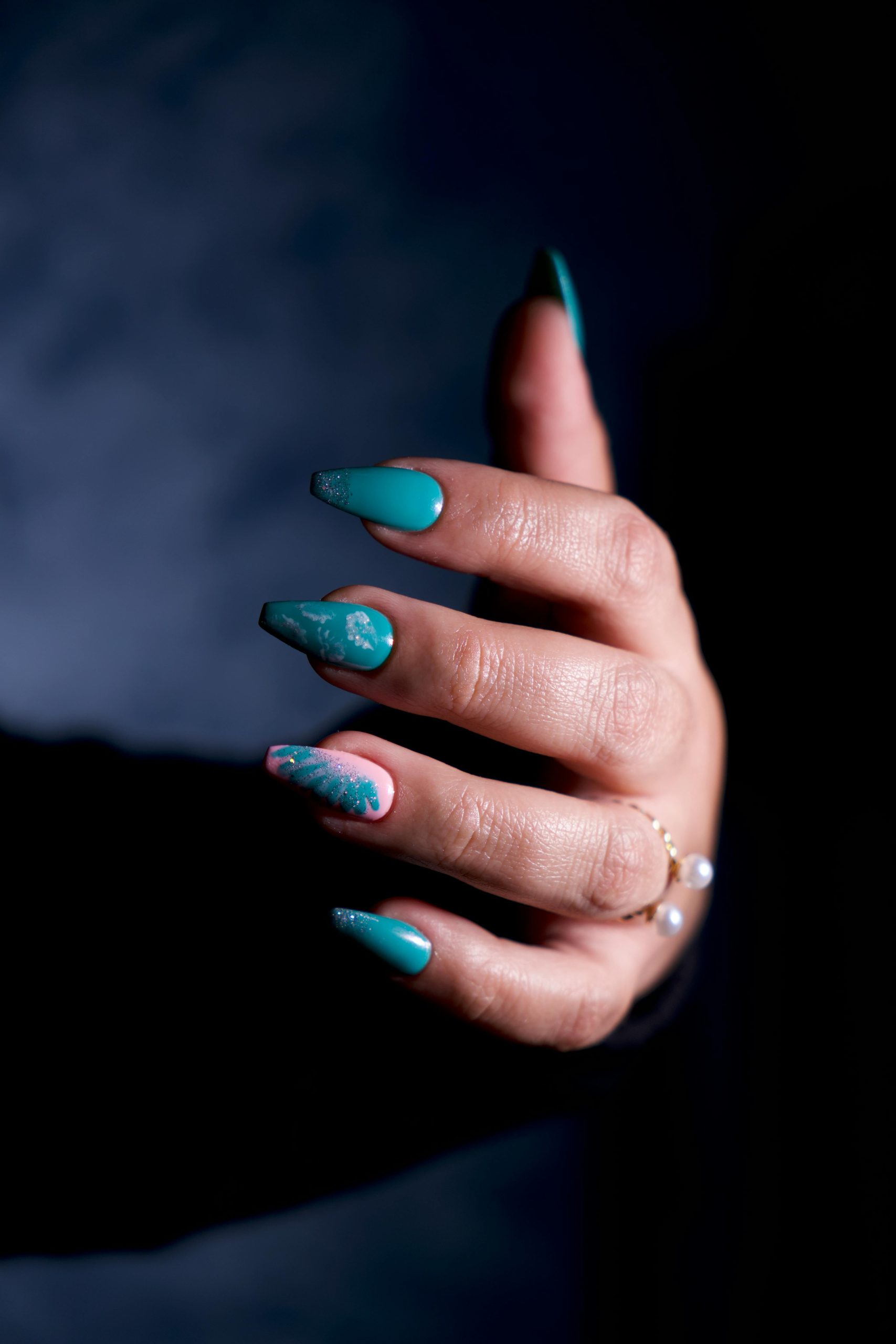 Elegant hand with turquoise nail art against a dark background, showcasing intricate designs. Main élégante avec nail art turquoise sur fond sombre, mettant en évidence des designs complexes.