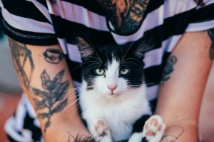 A close-up of a tattooed individual holding a black and white cat, both looking directly at the camera. Gros plan d'une personne tatouée tenant un chat noir et blanc, tous deux regardant directement vers l'appareil photo.
