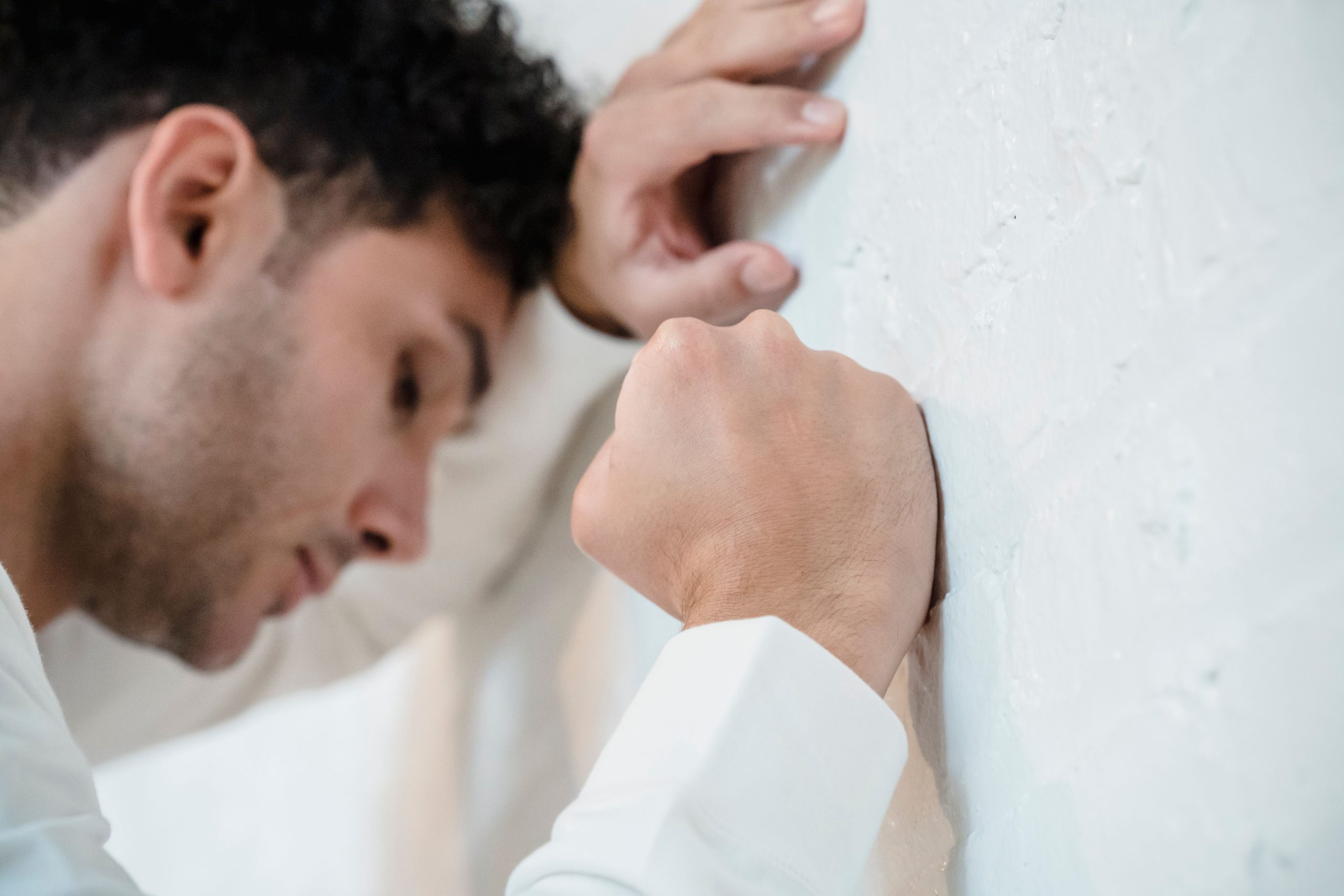 A man in distress leans on a white wall, depicting stress and anxiety. Un homme en détresse s'appuie contre un mur blanc, dépeignant le stress et l'anxiété.