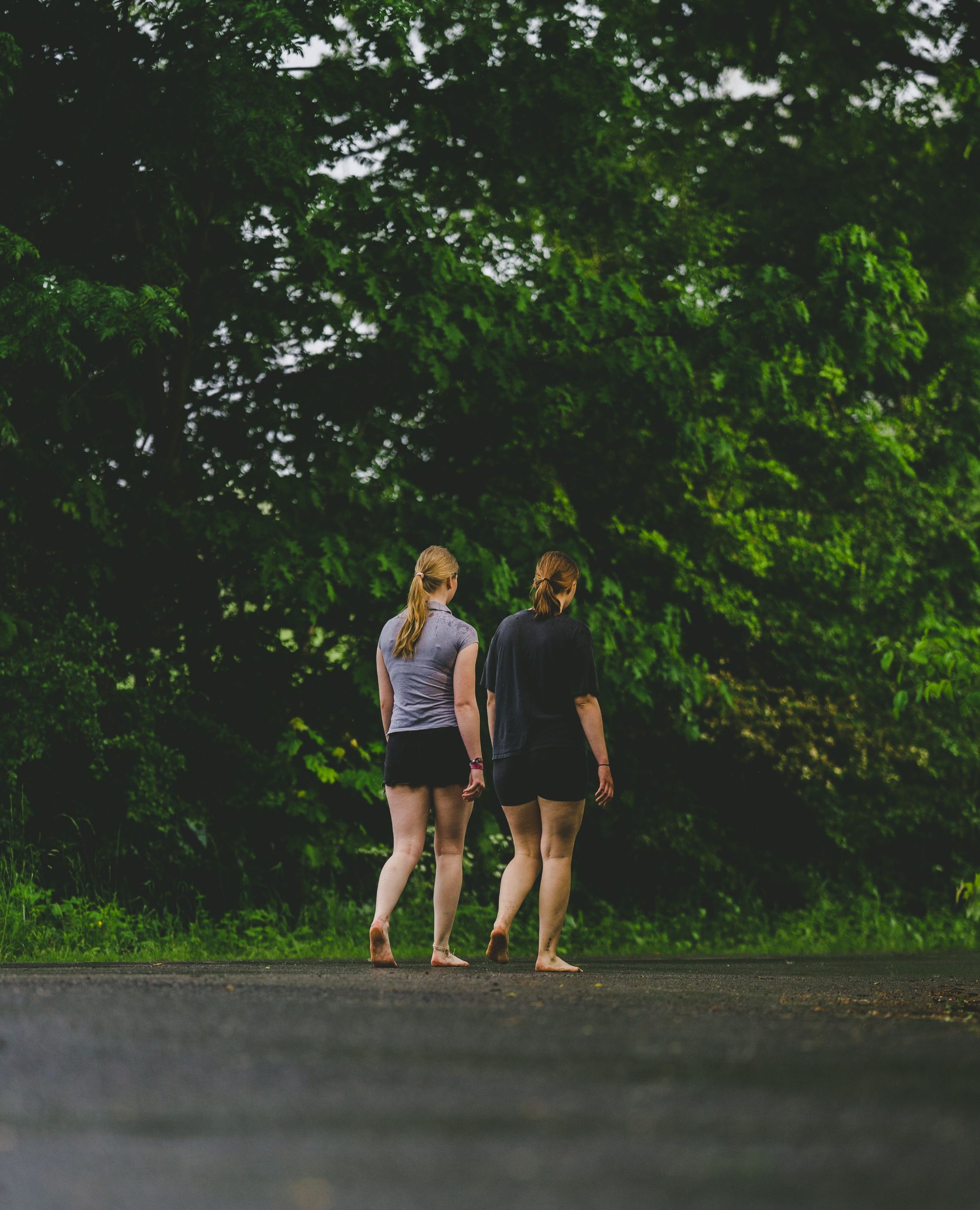 Two women walk barefoot on a road surrounded by green trees, showcasing tranquility and nature. Deux femmes marchent pieds nus sur une route entourée d'arbres verts, mettant en avant la tranquillité et la nature.