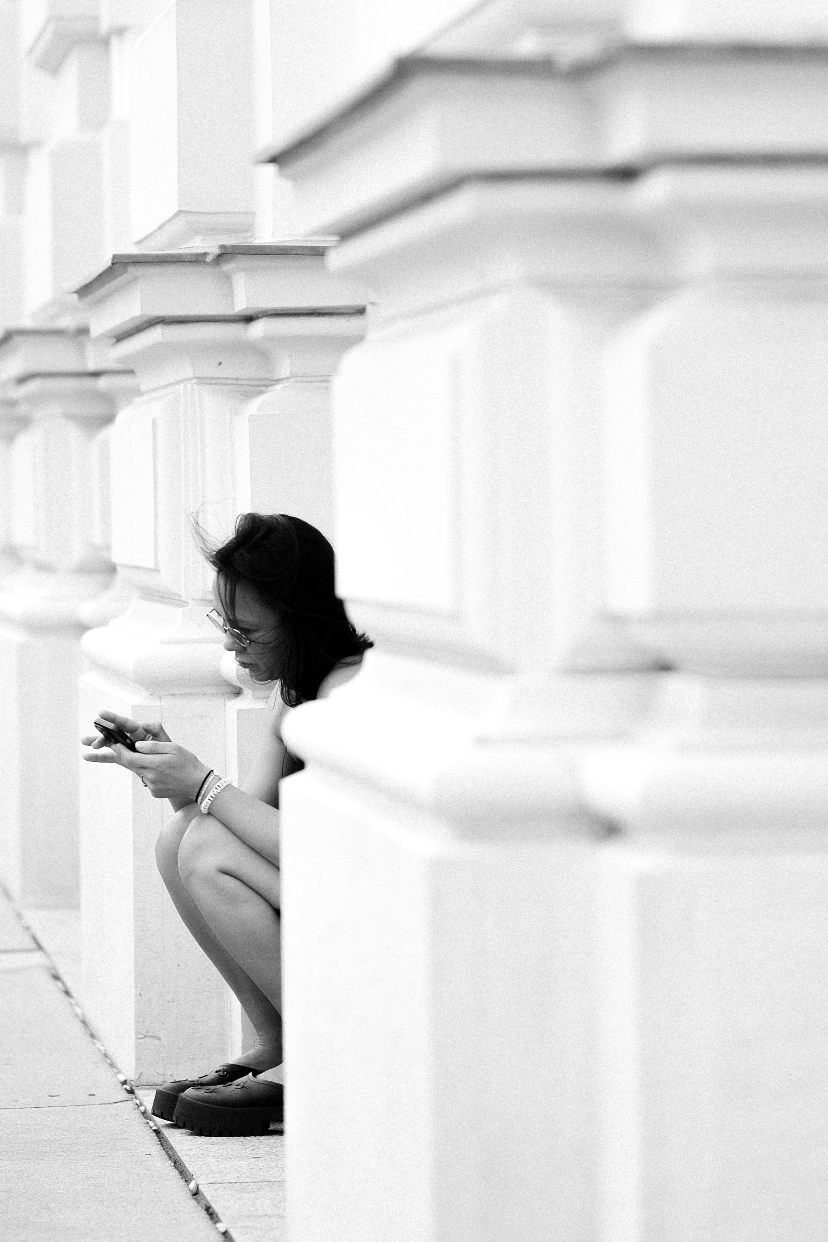 A woman crouched by a column in black and white, checking her phone. Une femme accroupie près d'une colonne en noir et blanc, vérifiant son téléphone.