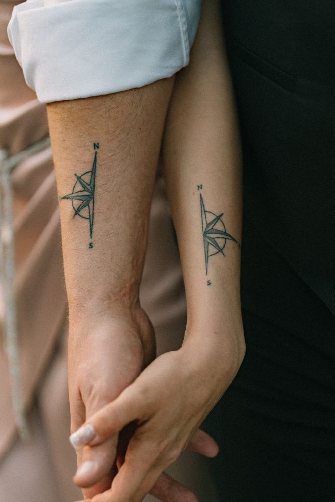 Close-up of a couple holding hands, showcasing matching compass tattoos on their forearms. Gros plan d'un couple se tenant la main, mettant en avant des tatouages de boussole assortis sur leurs avant-bras.
